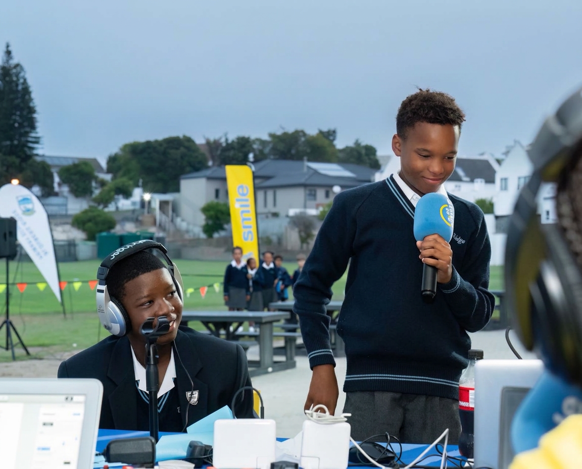 Two schoolchildren at an outdoor radio broadcast, one listening with headphones at a laptop and the other holding a microphone